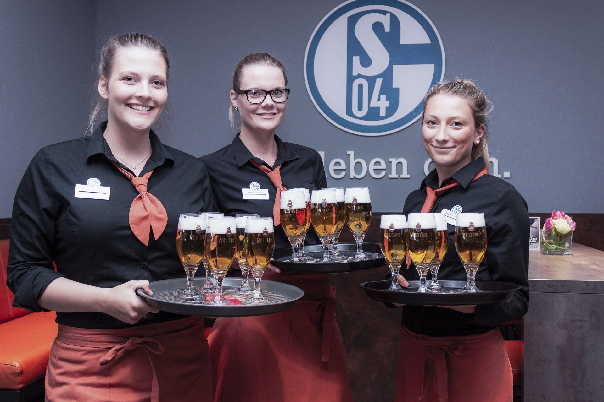 Drei lächelnde Frauen in Uniform mit orangefarbenen Krawatten halten Tabletts mit Biergläsern. Sie stehen in einem Innenraum in der Nähe einer Wand mit einem großen S04-Logo. Im Hintergrund ist ein Tisch mit Blumen zu sehen.