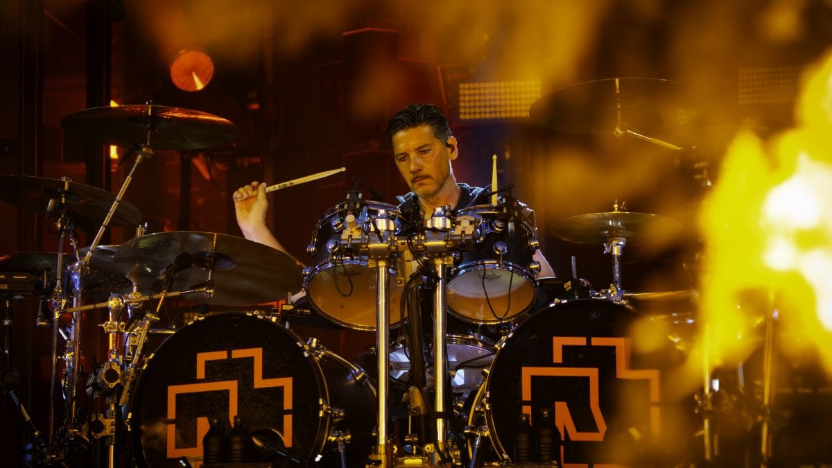Schlagzeuger spielt auf einer Bühne mit zwei Bassdrums, die das Rammstein-Logo zeigen, umgeben von gelbem Bühnenlicht
