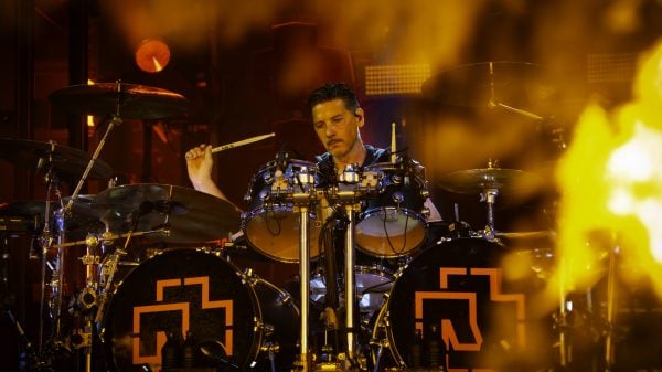 Schlagzeuger spielt auf einer Bühne mit zwei Bassdrums, die das Rammstein-Logo zeigen, umgeben von gelbem Bühnenlicht