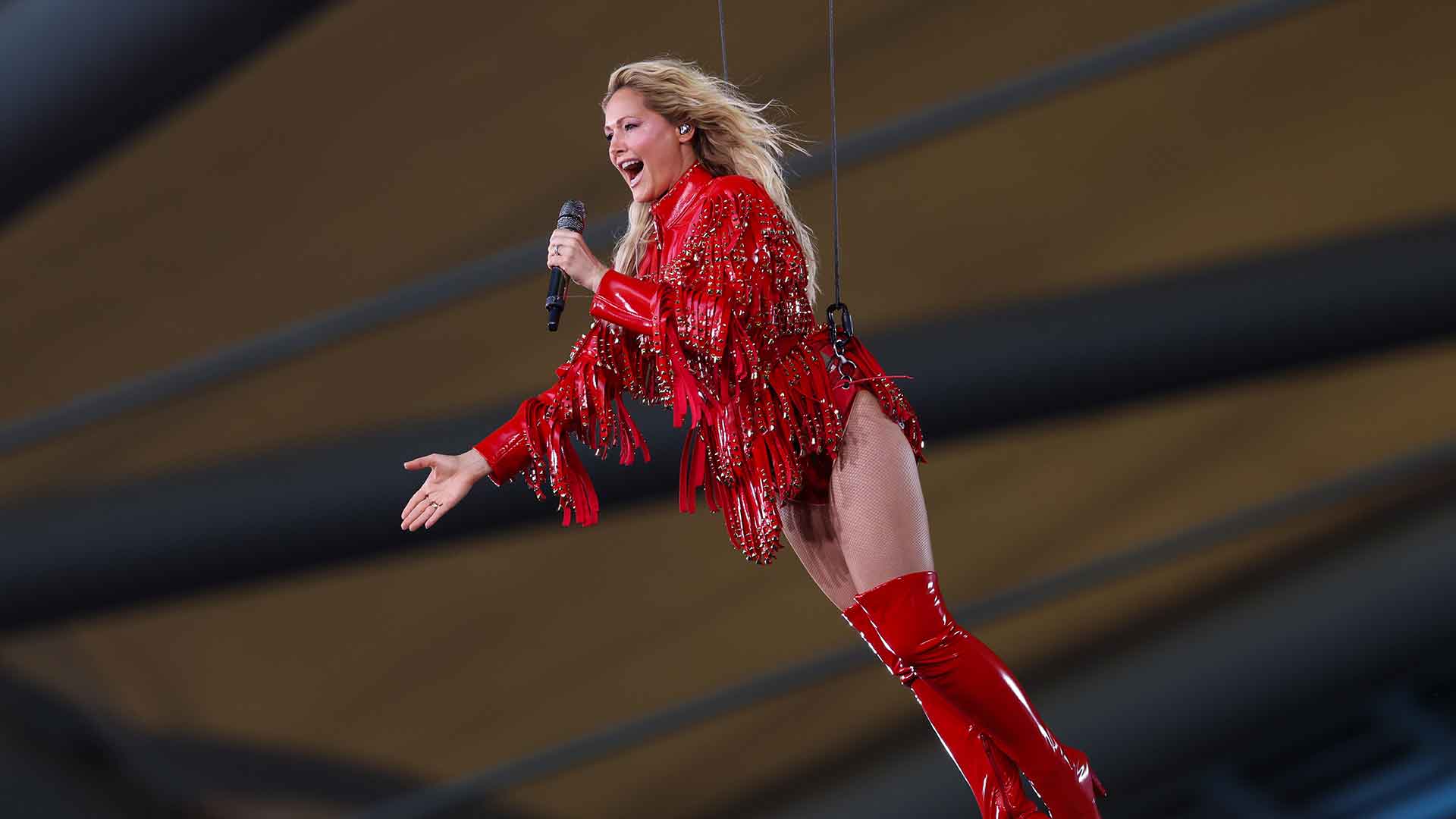 Helene Fischer in rotem Glitzer-Outfit und Overknee-Stiefeln, am Seil unter dem Arenadach schwebend, Mikrofon in der Hand und Arm ausgestreckt.